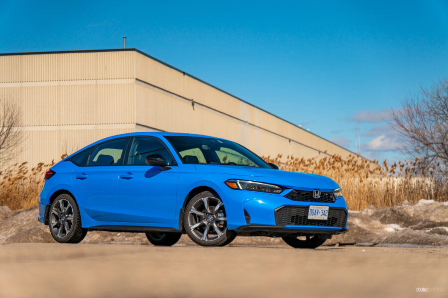Front quarter view of a blue 2025 Honda Civic Hybrid Hatchback on a sunny day, with trees and a building in the background