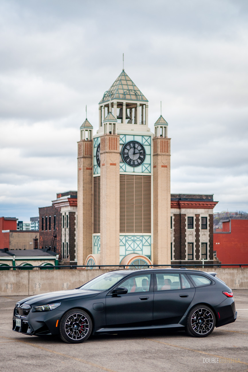Front quarter view of a satin black 2025 BMW M5 Touring in an empty parking lot on a cloudy day, with buildings in the background